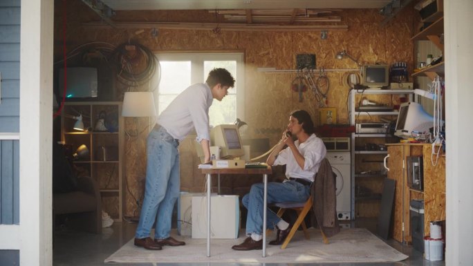 两个白人男性股票交易员在复古车库里工作，使用带有烛台图和固定电话的旧台式电脑向投资者推销一家上市公司
