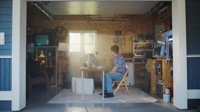 白人男性软件工程师走进复古车库，坐在他的办公桌前，旁边是一台旧台式电脑。专注于90年代创办软件创业公