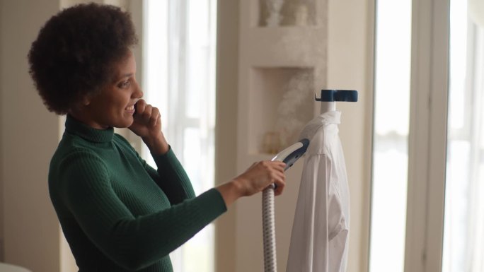 积极的非裔美国年轻女性的肖像，一边讲着智能手机，一边在家用手工蒸汽机蒸白衬衫。