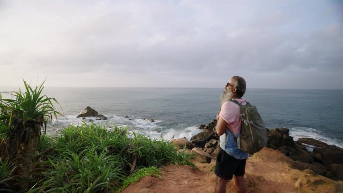 有灰胡子的老男人背着双肩包，从热带岛屿的海角眺望海洋、天空和充满异国情调的大自然。老年男性退休独自旅