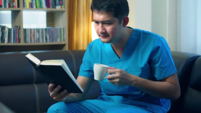 护士亚洲男工人喜欢看书，喝咖啡在家里的沙发上。