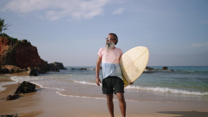 一位白发老人留下来，穿着衬衫，t恤，戴着太阳镜，在沙滩上摆着冲浪板的姿势。黑人老人微笑着面对镜头，在
