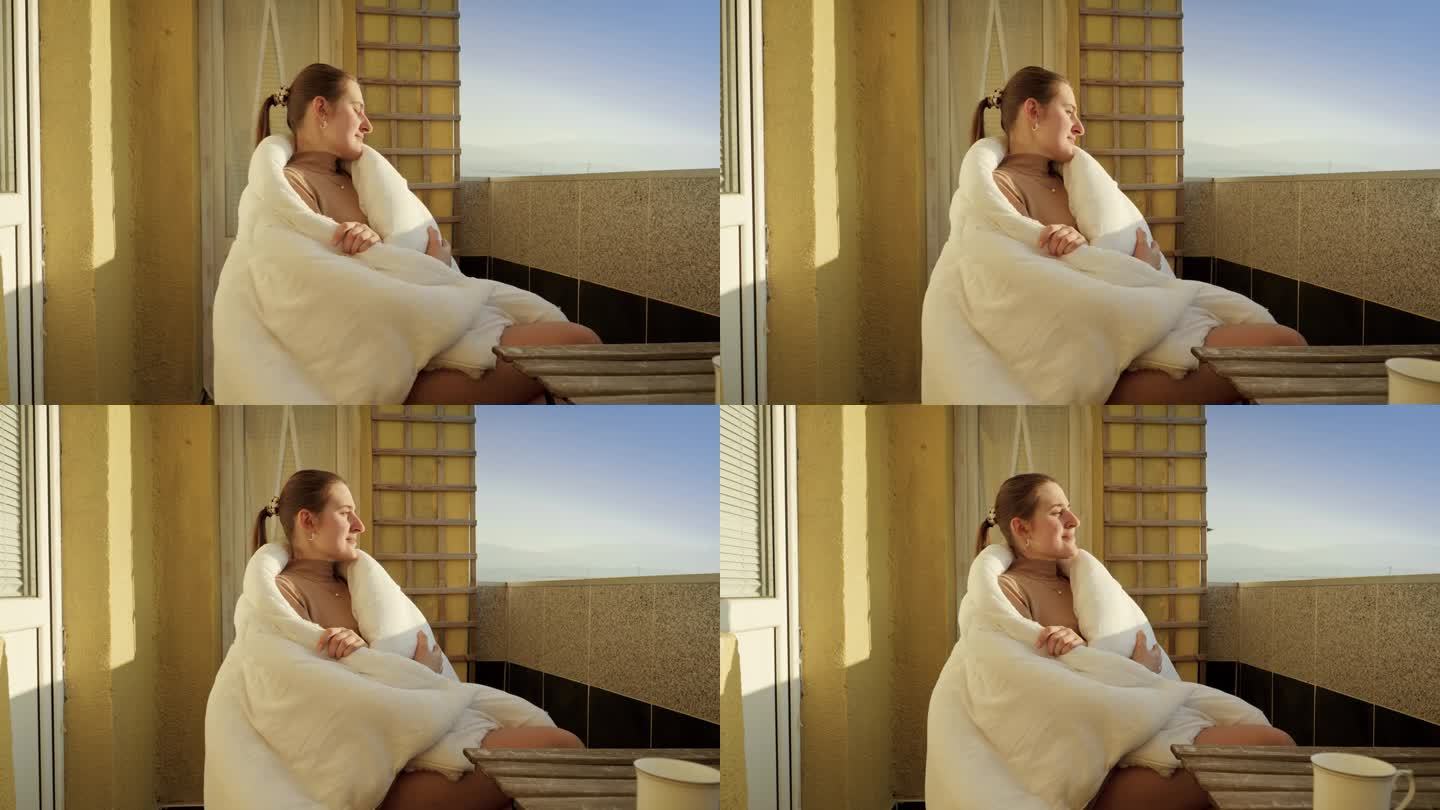 年轻女子披着毯子坐在阳台上看太阳。早晨的人们，在家里放松，欣赏城市美景