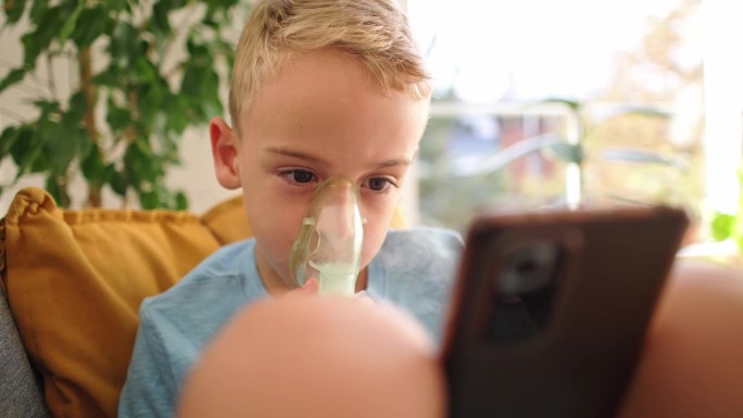 患有哮喘的高加索男孩，在治疗期间，一边使用雾化口罩，一边坐在沙发上用手机看卡通片