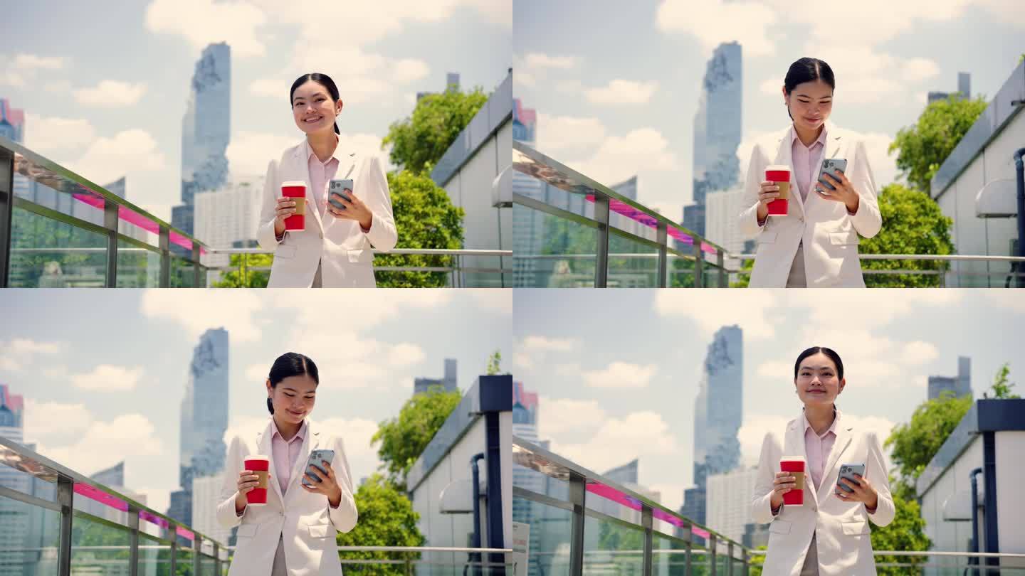 一位年轻的亚洲女商人拿着红色的咖啡杯，玩着智能手机，自信地走在绿色屋顶公园里。