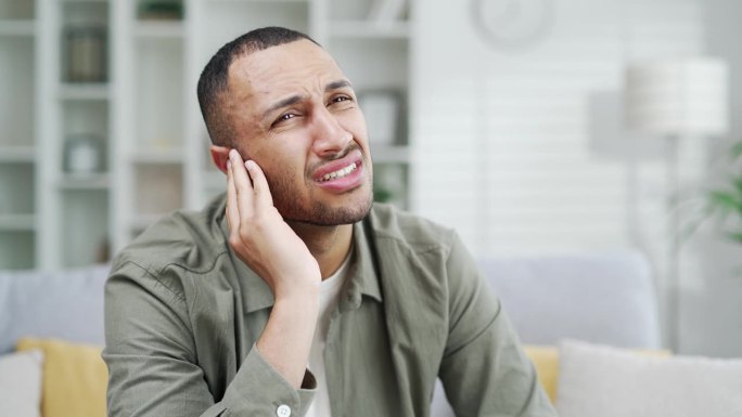 男人在家时耳朵疼。男性坐在客厅的沙发上抱着疮。中耳炎或室内发炎
