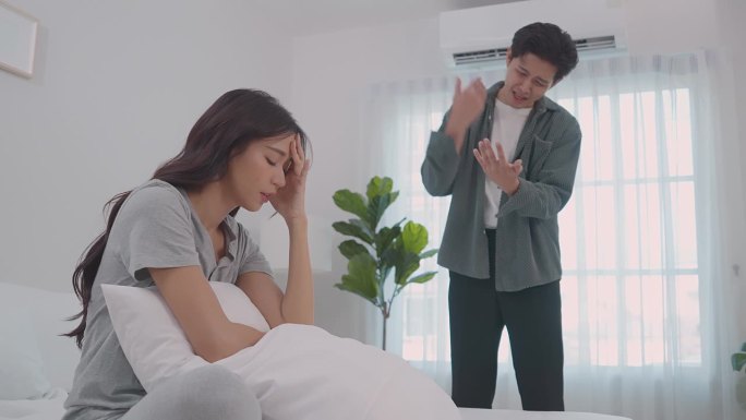 亚洲年轻夫妇坐在床上痛苦的争吵后打架。新婚男女因在卧室里吵架而伤心欲绝。紧张的亚洲女人坐在床上哭