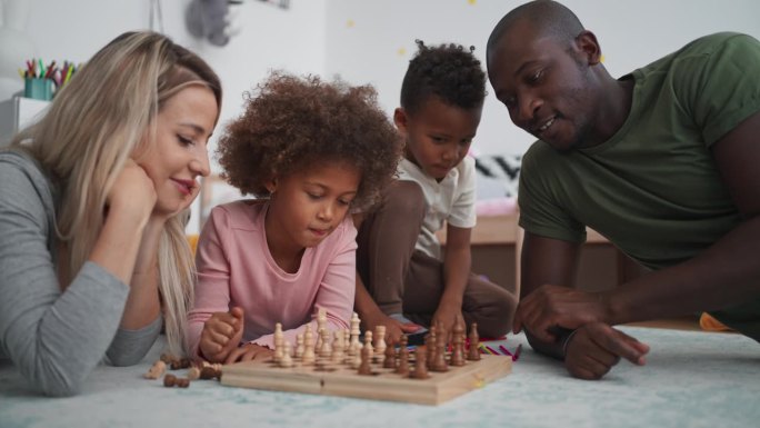 年轻的多种族家庭，两个小孩躺在地板上，在家里下棋。