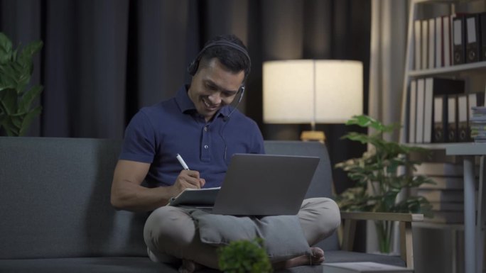 一个年轻的黑发男子坐在沙发上，用他的笔记本电脑工作。年轻男子远程工作男子戴着耳机看电影，听音乐在一个