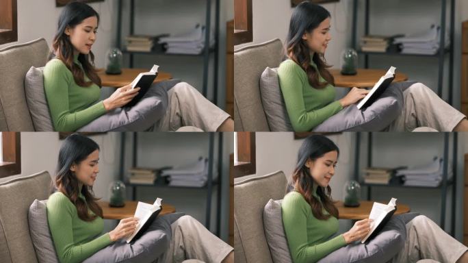 微笑的亚洲女人在家里看书，在沙发上放松
