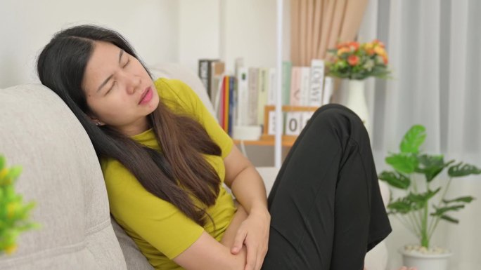 一名亚洲妇女胃痛严重，她感到压力很大，在家里的沙发上痛苦不堪。