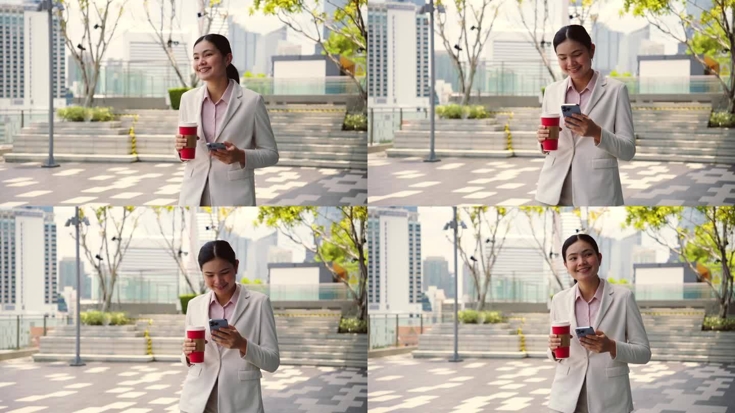 一位年轻的亚洲女商人拿着红色的咖啡杯，玩着智能手机，自信地走在绿色屋顶公园里。