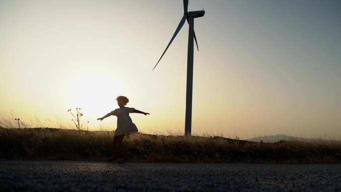 快乐的孩子女孩的剪影旋转，转身双手在大自然的夕阳在田间与风车风力发电机。自由，童年，环保能源动力。