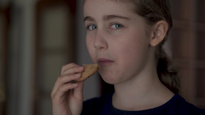 吃饼干的小女孩。看着镜头，微笑着。可爱的孩子饥饿吃饼干享受美味的治疗在家里的厨房。垃圾食品，不健康的