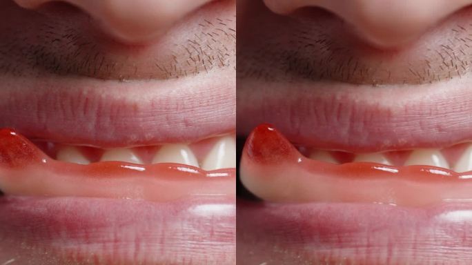 垂直视频。一个留着胡子的年轻男子嘴里叼着像红色吸血鬼尖牙一样的软糖吃着。