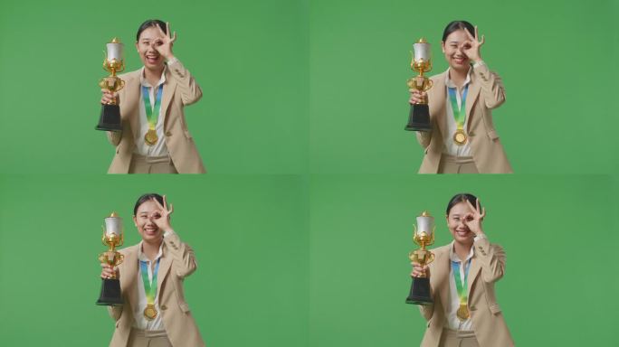 亚洲女商人穿着西装，拿着金牌和奖杯，在绿幕背景下对着镜头微笑，成为第一位获奖者