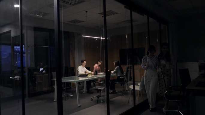 三名员工在正常工作时间之外加班，办公室里只有会议室的灯还亮着。