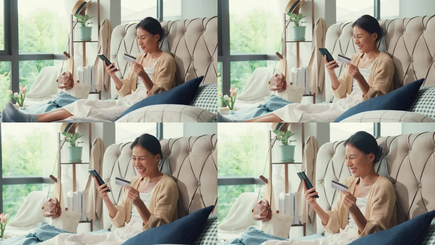 快乐的亚洲女孩大学穿着白色的奶油睡衣躺在柔软舒适的床上使用信用卡网上购物成功的手机支付早上在家里的卧