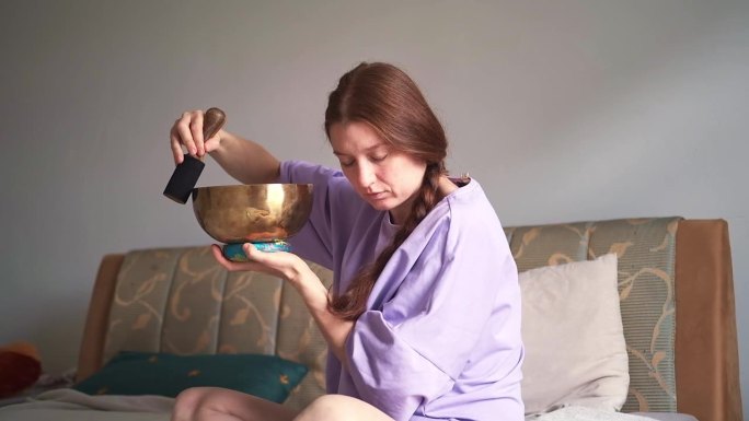 一个红头发的女人正坐在家里的床上，拿着唱歌的碗打坐。