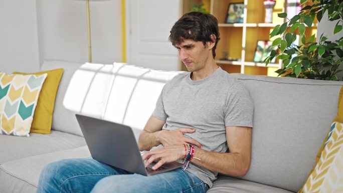 年轻的西班牙人坐在沙发上使用笔记本电脑，在家忍受胃痛