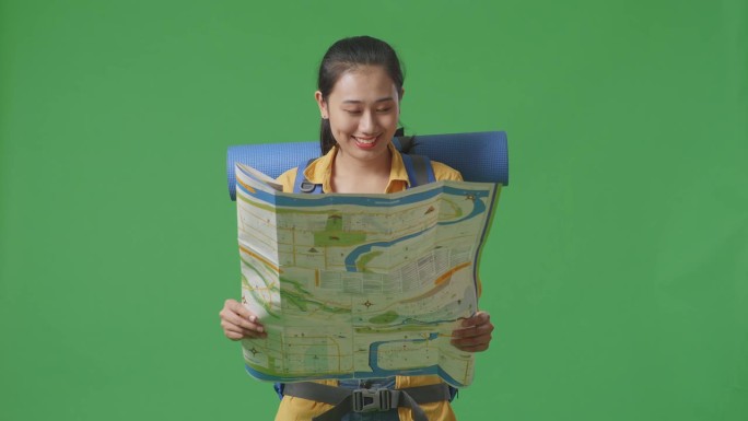 扛着登山背包的亚洲女徒步旅行者站在摄影棚的绿幕背景上，看着地图，然后对着镜头微笑