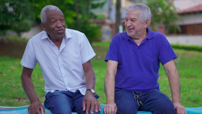 一位非裔美国老人坐在一位白人朋友旁边享受着退休后的户外公园，两个不同的老人之间的互动
