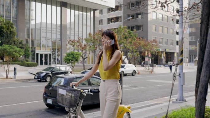 一名亚洲女子推着一辆共享单车，在城市街道上用智能手机与同事或朋友交流。