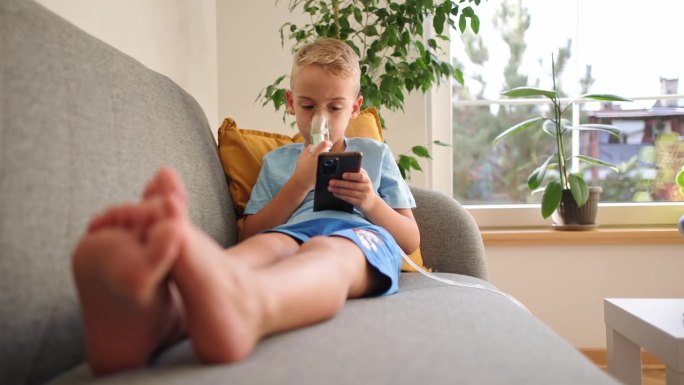 患有哮喘的高加索男孩，在治疗期间，一边使用雾化口罩，一边坐在沙发上玩手机视频游戏