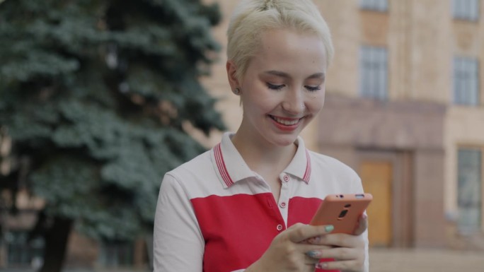 无忧无虑的年轻女学生用智能手机查看社交媒体，站在城市外面笑着