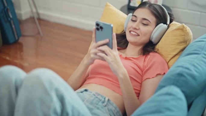 幸福的女人戴着耳机，用智能手机躺在家里的沙发上