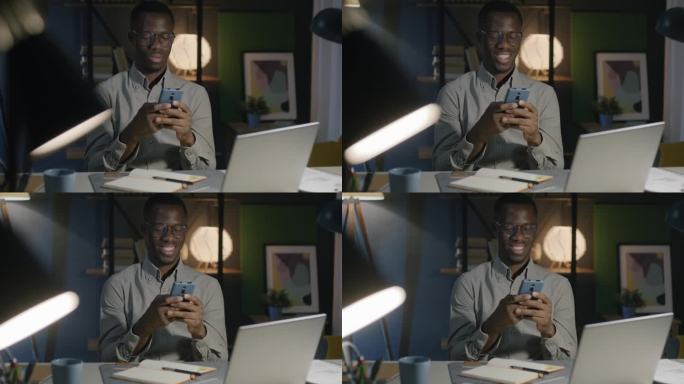 非裔美国人自由职业者，晚上在家用智能手机和客户聊天，微笑着工作