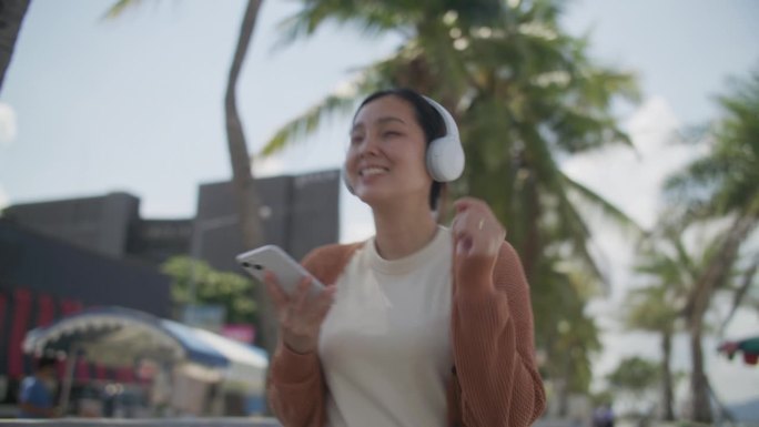 年轻的亚洲女子戴着无线耳机，拿着智能手机在沙滩上跳舞