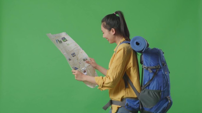侧视图的亚洲女性徒步旅行者与登山背包看地图，然后环顾四周，而走在绿屏背景在工作室