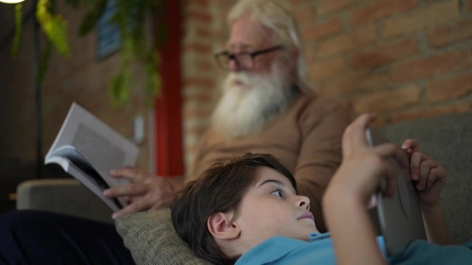 爷爷在看书，孙子在家里玩数码平板电脑