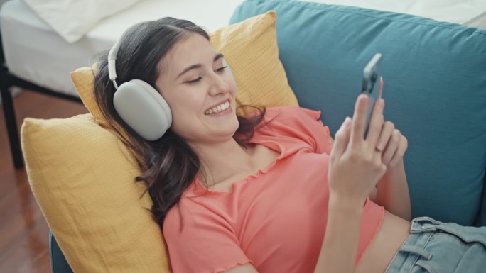 幸福的女人戴着耳机，用智能手机躺在家里的沙发上