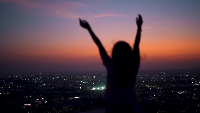 一个十几岁的女孩在落日的余晖中举手，背景是这座城市的剪影。欣赏城市夜景的女游客。