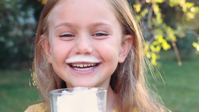 快乐的情绪微笑着可爱的孩子女孩拿着一杯牛奶，喝在阳光明媚的夏日早晨的大自然。孩子张开嘴，嘴唇上有牛奶