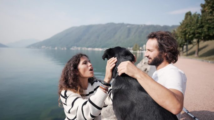 男人和女人在湖边和他们的狗玩耍，微笑着