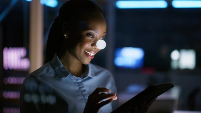 快乐的黑人女性晚上在办公室里摆弄着平板电脑，为社交媒体进行网络、研究和营销规划。迟到，在工作场所阅读