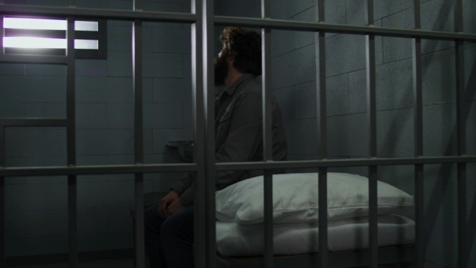 罪犯坐在牢房的床上，望着铁窗