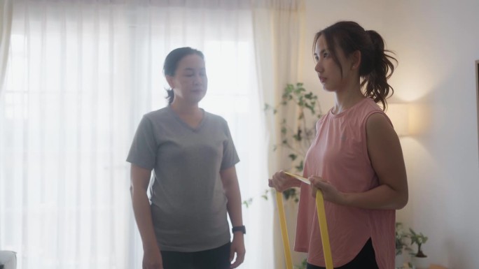 私人教练向亚洲老年女性展示如何在家中客厅使用阻力拉伸带运动。