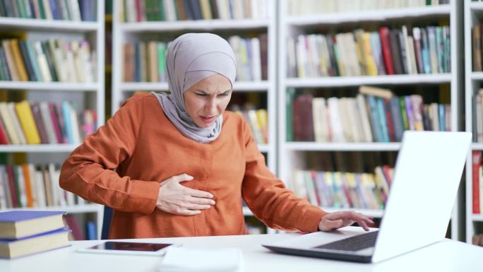 戴着头巾的穆斯林女学生在校园图书馆用笔记本电脑工作时胃痛