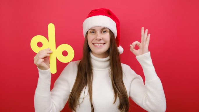 可爱有趣的年轻女子穿着温暖的白色毛衣，戴着圣诞老人帽，手里拿着大大的黄色标记，在柔和的红色工作室背景