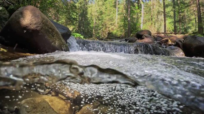 相机缓缓跳入高山河流中最纯净的充满气泡的水中。半水下慢动作拍摄的河流在绿色的森林和山脉。石头之间有小