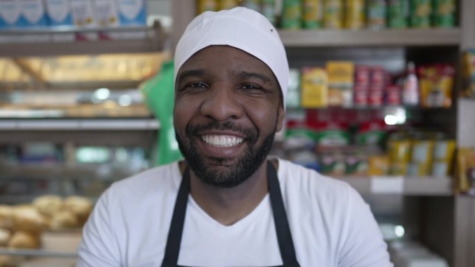 在自助餐厅柜台后面快乐的非裔美国工人，穿着制服，产品和消费品在背景中模糊，对着镜头微笑