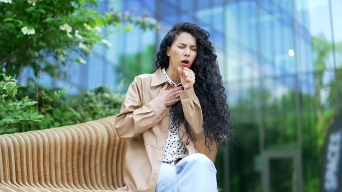 生病的年轻女员工坐在办公大楼附近街道的长凳上咳嗽。疲惫的女人把手放在胸前