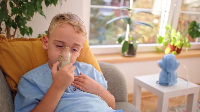 疲惫的哮喘高加索男孩，坐在沙发上使用雾化口罩，在家庭治疗期间