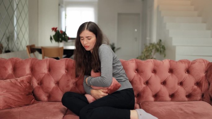 患病的年轻女子遭受经期疼痛。坐在沙发上有腹痛、胃炎、痛经等症状的手捏肚子的女性。