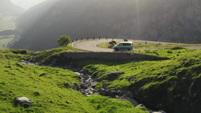 银色露营车沿着蜿蜒的瑞士公路弯道行驶。清晨。牛在弯道里吃草。形象生动，草木生机勃勃，山涧淙淙。电影般