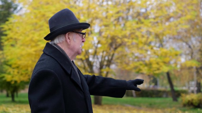 一位爷爷走过一个美丽的秋天公园，他戴着一顶黑帽子，穿着大衣，讲述着他童年的回忆。
